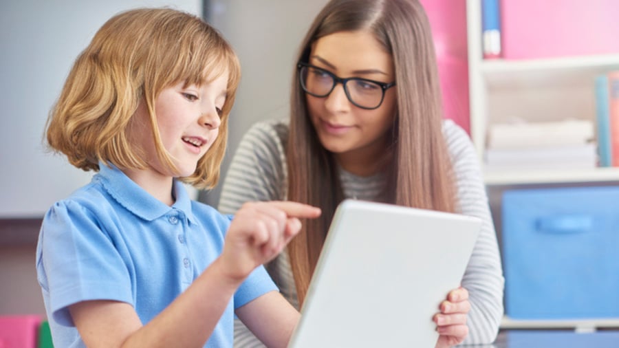 a young teacher listens as her young pupil explains what she is reading on a digital tablet . 